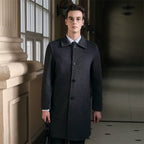 Male model wearing a dark gray men's pure wool slim fit overcoat with a classic collar, button closure, and tailored fit, standing indoors near a window