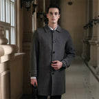 Men's slim fit overcoat in dark gray wool fabric, classic collar, button closure, worn by a young man in an elegant indoor setting